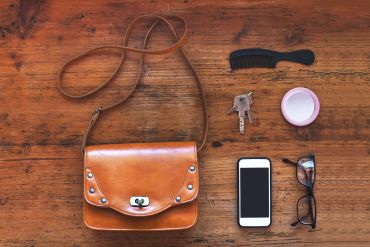 Flat lay of everyday essentials including a brown leather bag, phone, keys, and glasses.