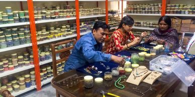 Three people crafting jewelry with beads in a workshop filled with jars of materials.